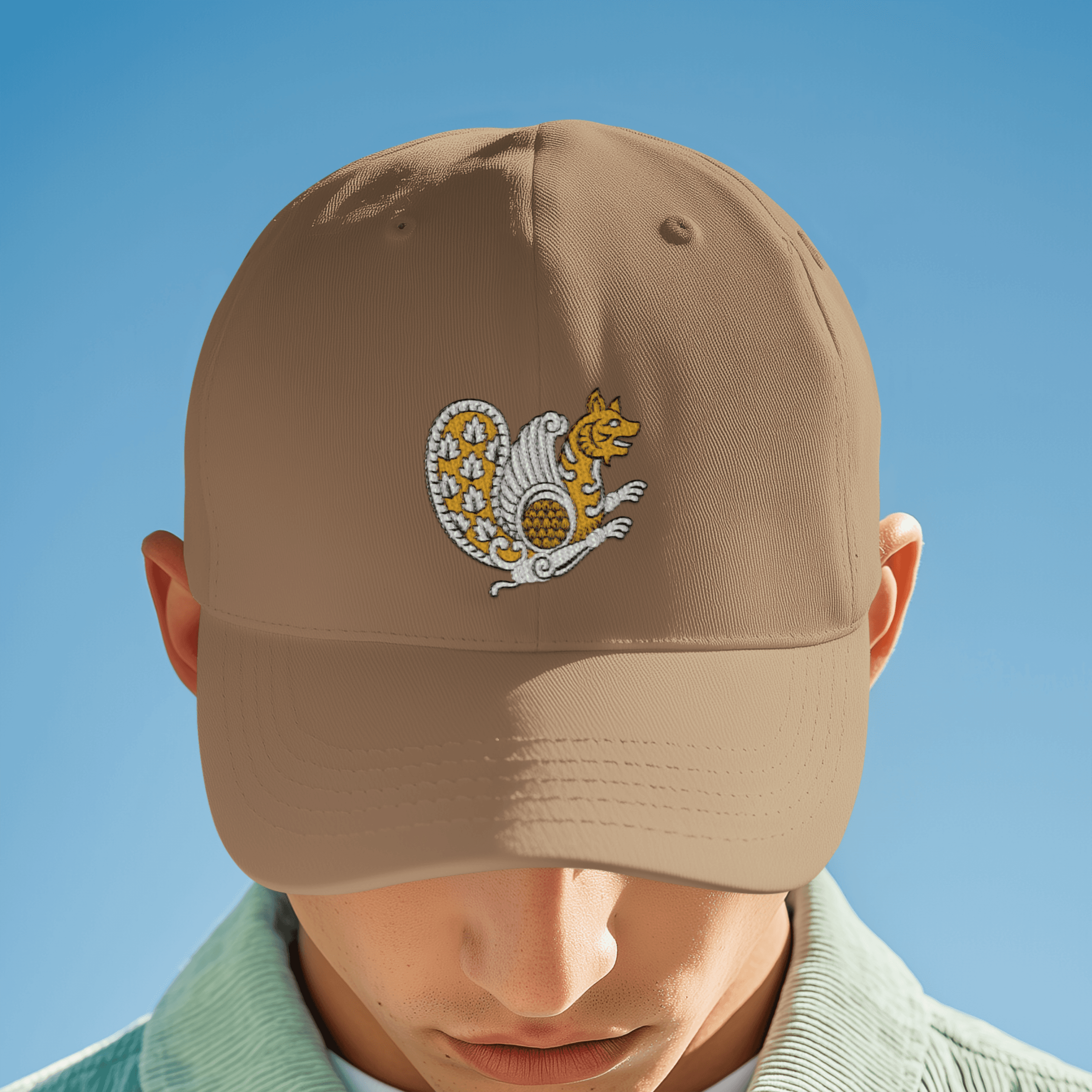 Front view of a tan dad cap embroidered with a stylized Simurgh bird symbol from Iran, photographed against a clear blue sky with the brim facing forward.