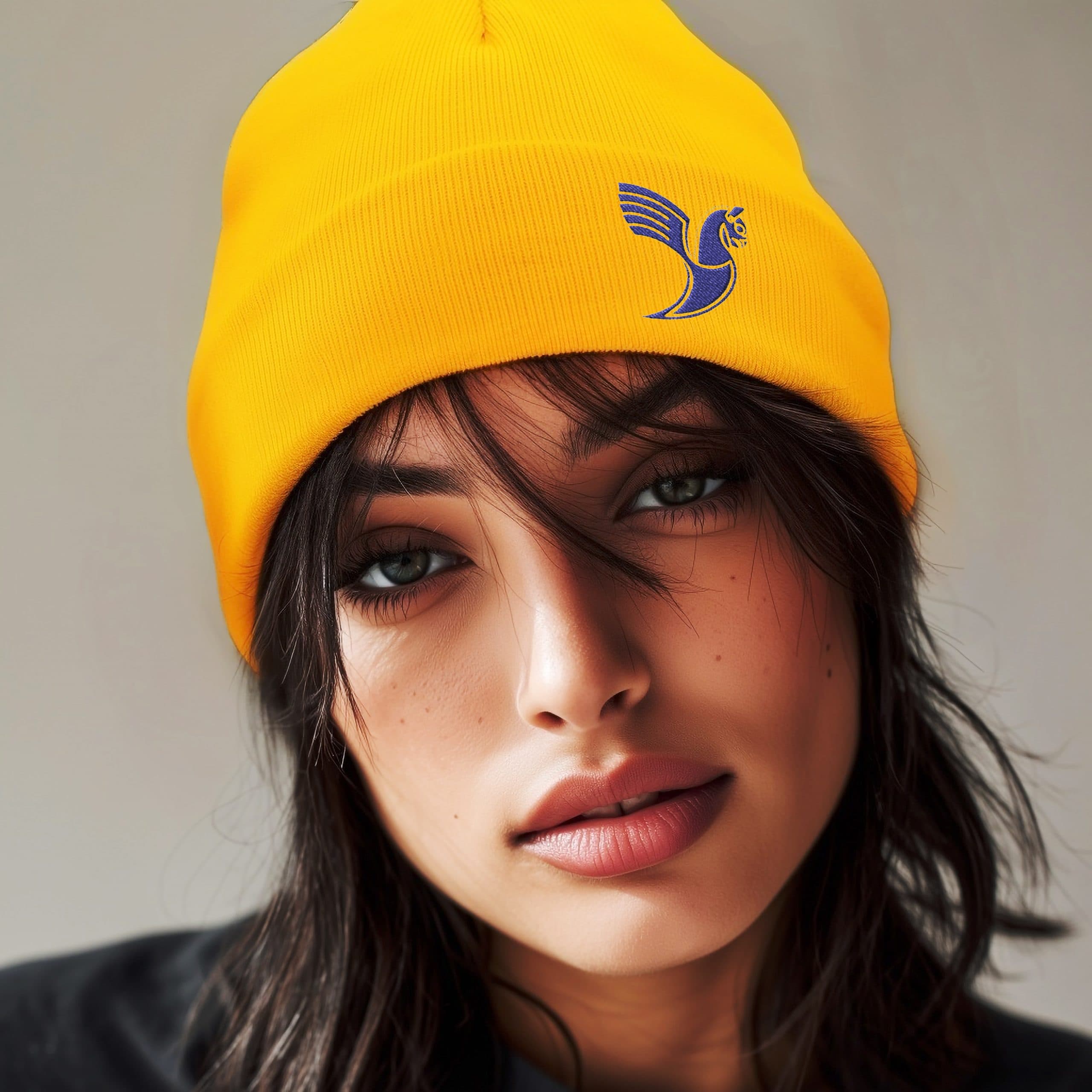 Close portrait of a woman wearing a yellow cuffed beanie with blue Huma bird embroidery, styled against a warm-toned background.