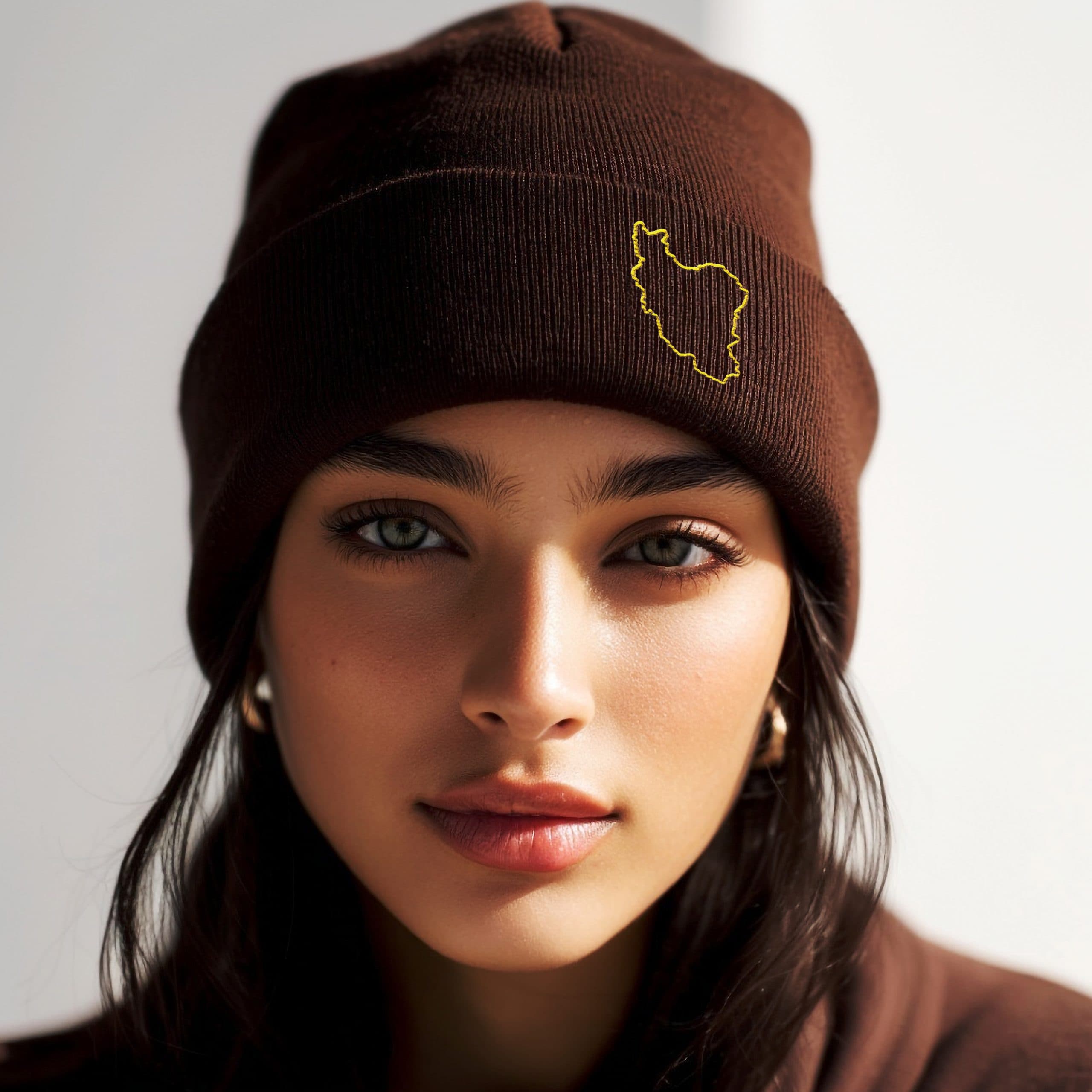 Woman wearing a brown cuffed beanie embroidered with a minimalist gold outline of Iran, photographed in a clean studio setting.